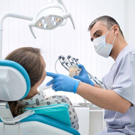 Dentist in mask and gloves shows a dental model to a young woman seated in a dental chair under overhead light