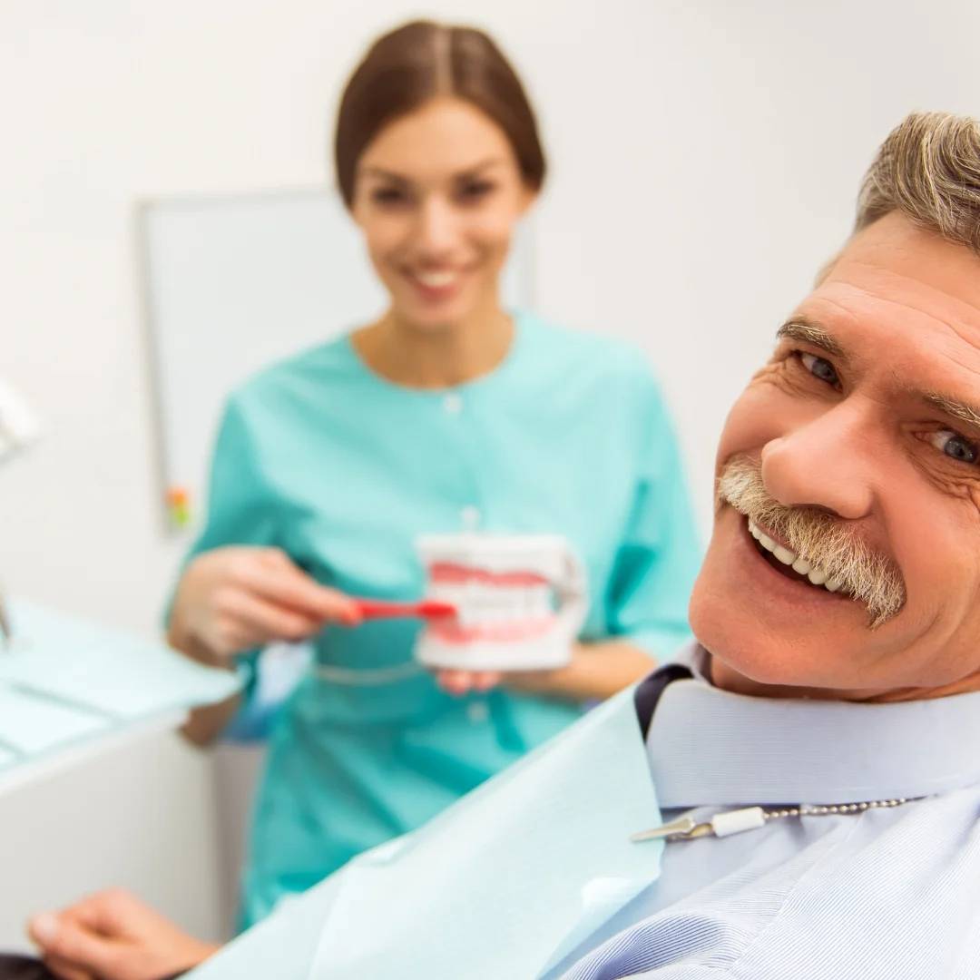Middle-aged man smiling while a dental professional holds a tooth model during a consultation